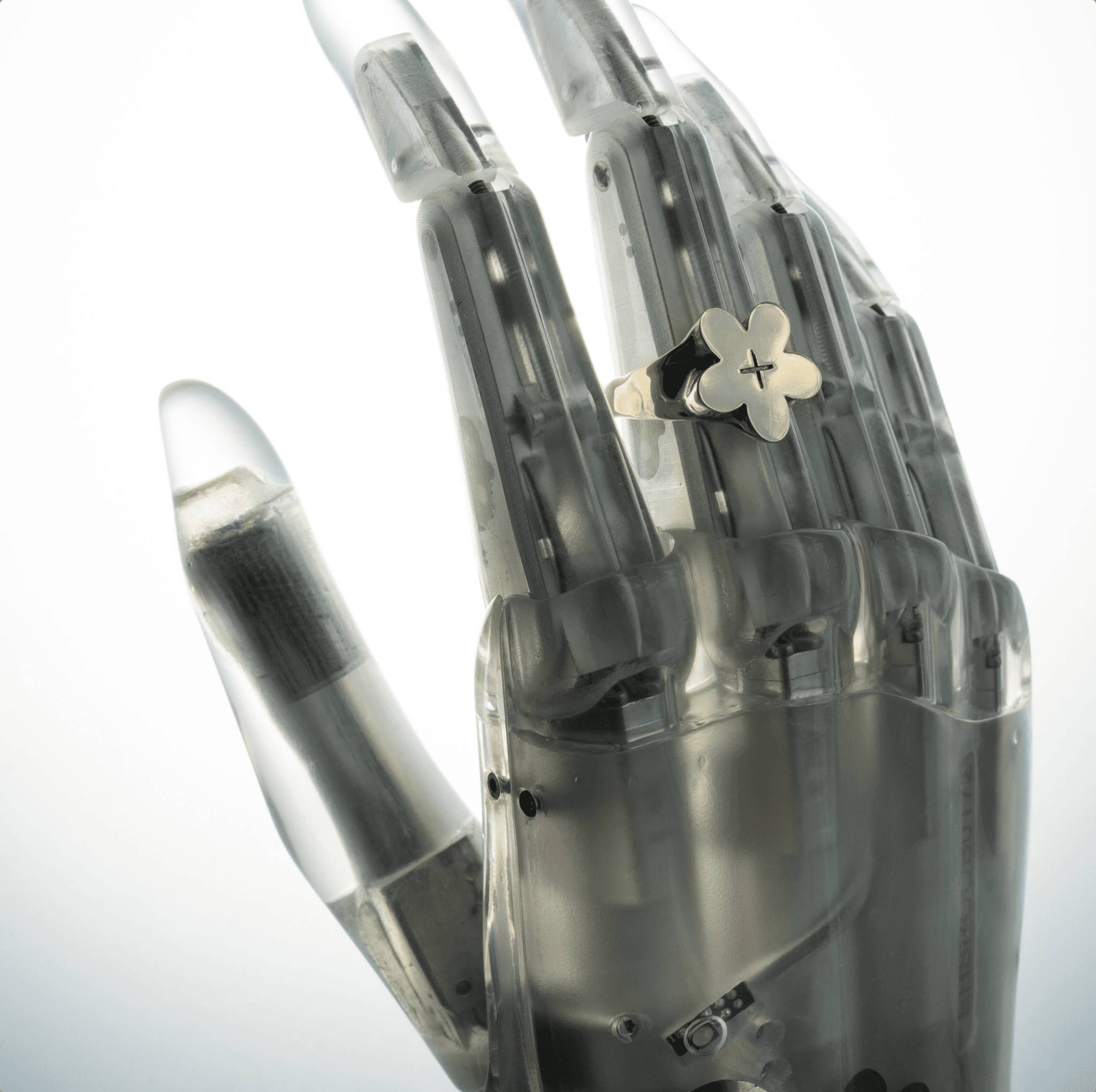 A close-up view of transparent prosthetic fingers made of lightweight aluminum and polycarbonate, revealing internal mechanisms. One finger holds a small gold flower ring, emphasizing strength and precision.