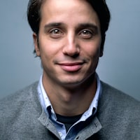 A headshot portrait of a young adult man with short brown hair, wearing a light blue collared shirt under a gray cardigan, against a plain blue-gray background. He has a slight smile and is looking directly at the camera.