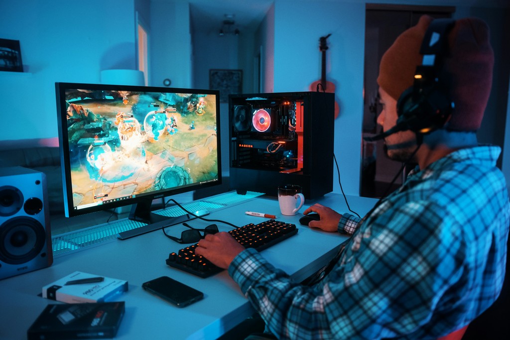 A dedicated gamer immersed in an online video game, wearing a headset and seated at a desk with a high-performance gaming PC, dual monitors, and ambient blue lighting in a cozy, modern room.