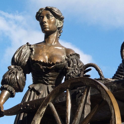 A bronze statue of a woman with a determined expression, standing next to a wagon wheel against a blue sky backdrop.