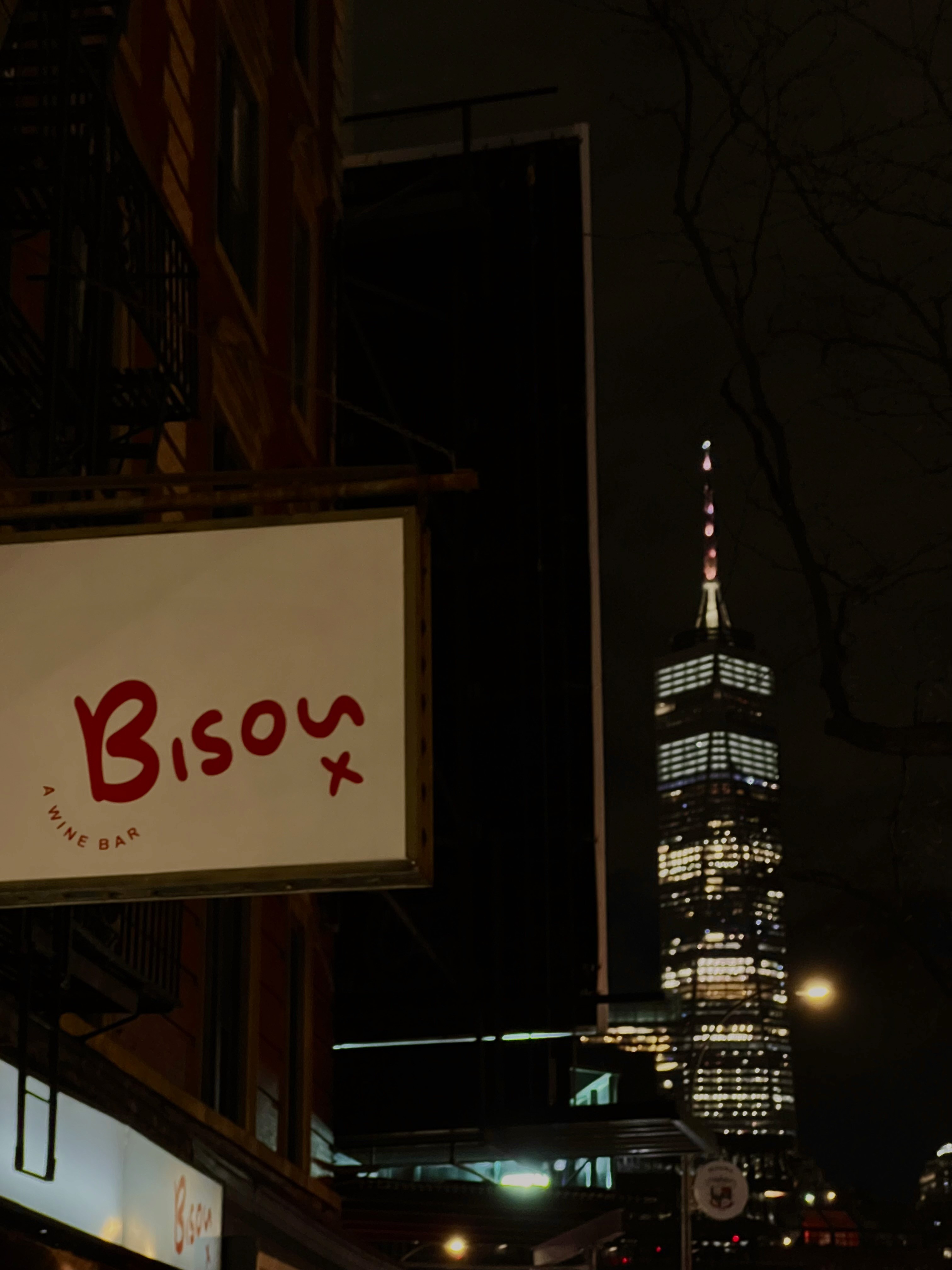 a sidewalk view of the new york skyline with a restaurant sign on the left that says "bisou" in red text