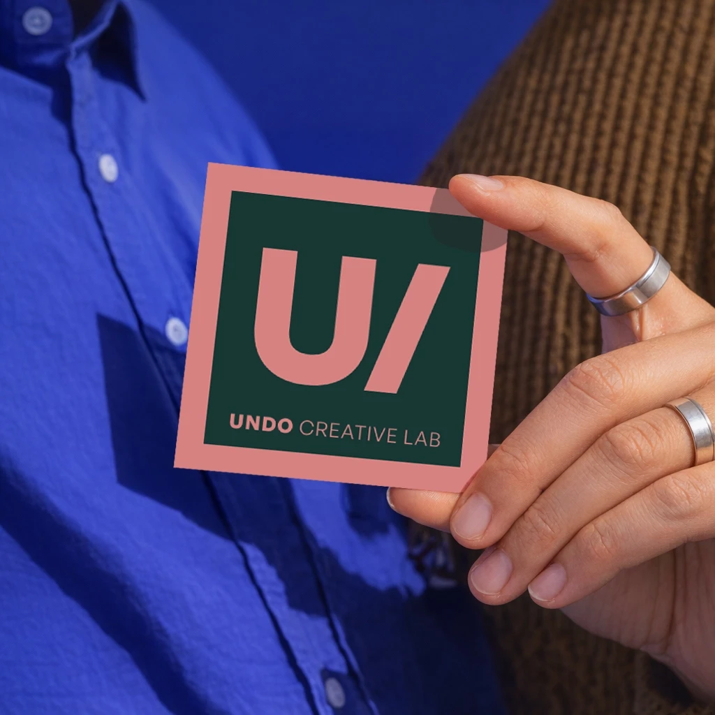 Hand holding Undo Creative Lab branded business card with green and pink logo design, person wearing blue shirt in background