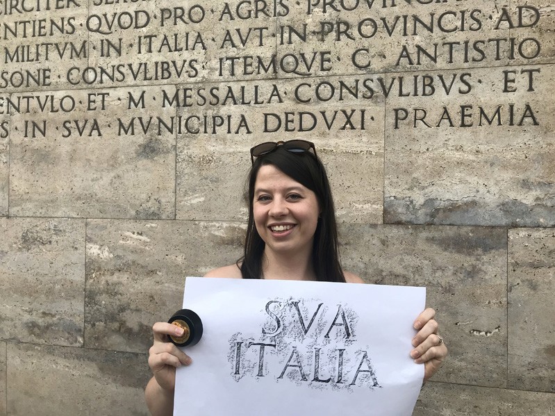 A photo of a girl holding a piece of paper with letters copied from a stone carved wall behind her.