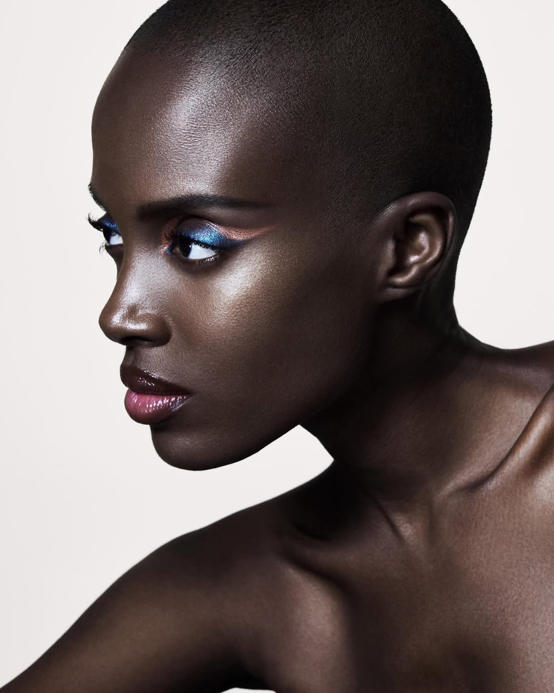 Model with shaved head wearing bold blue and pink eye makeup, looking to the side under dramatic lighting for Armani Beauty