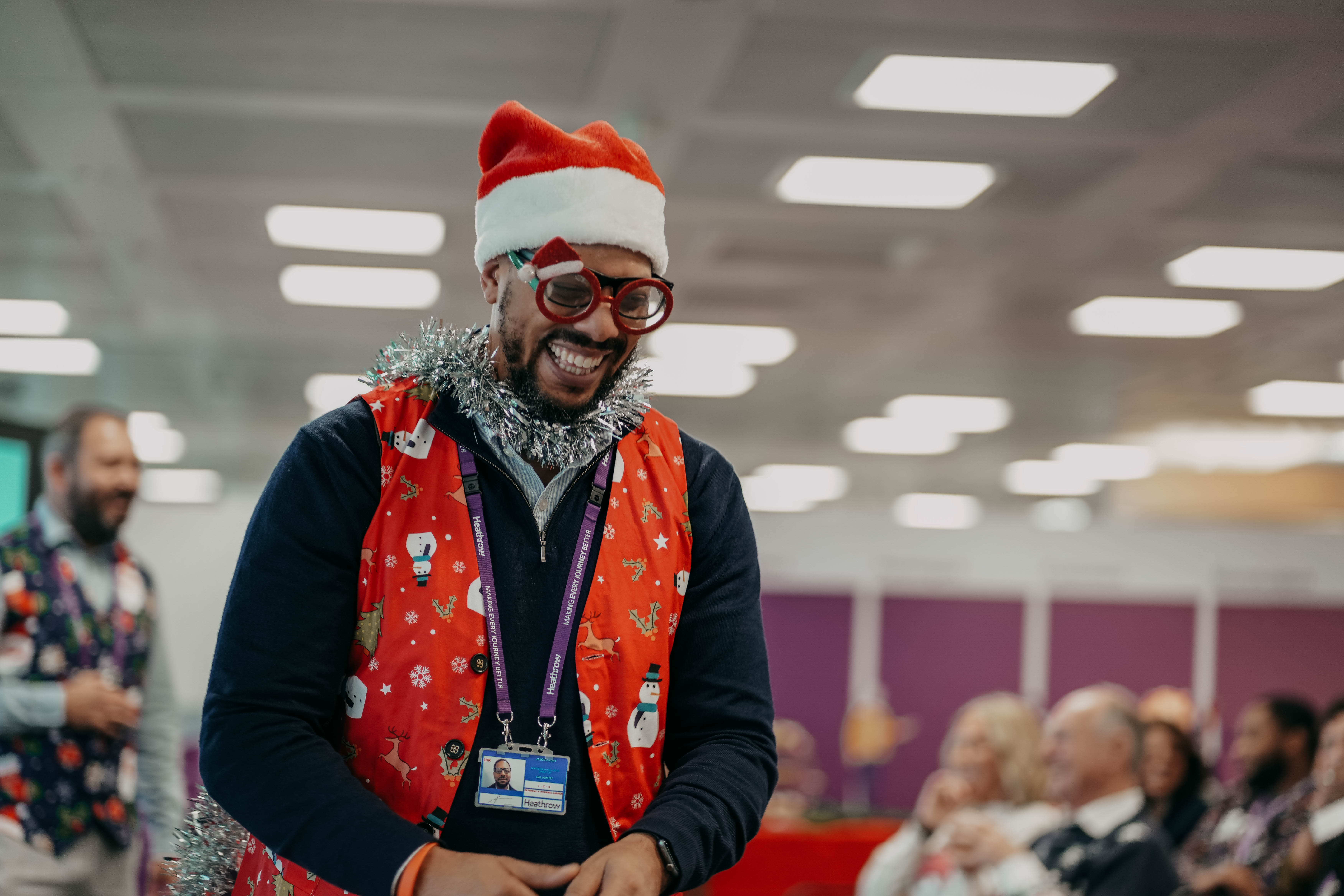 A man laughs heartily at an office Christmas party, wearing a Santa hat, novelty glasses, a red festive vest over his shirt, and silver tinsel around his neck.