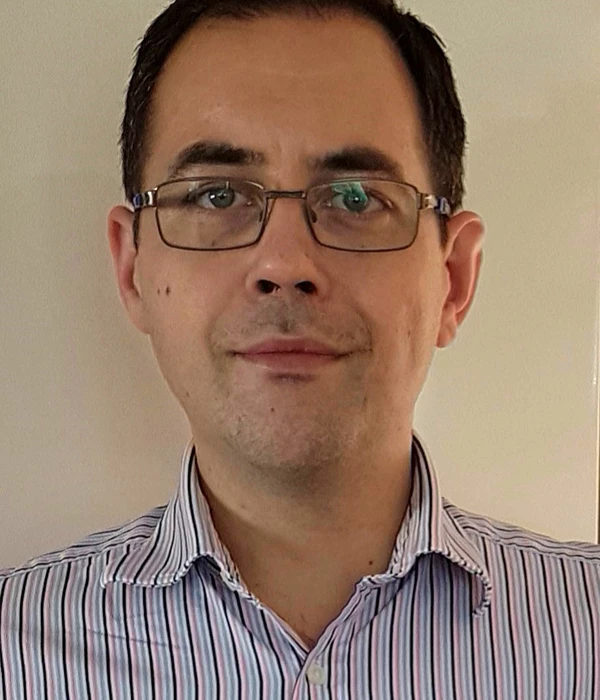 A person wearing a light‑coloured, striped button‑down shirt stands against a plain indoor wall. The framing and neutral background suggest a simple professional headshot.
