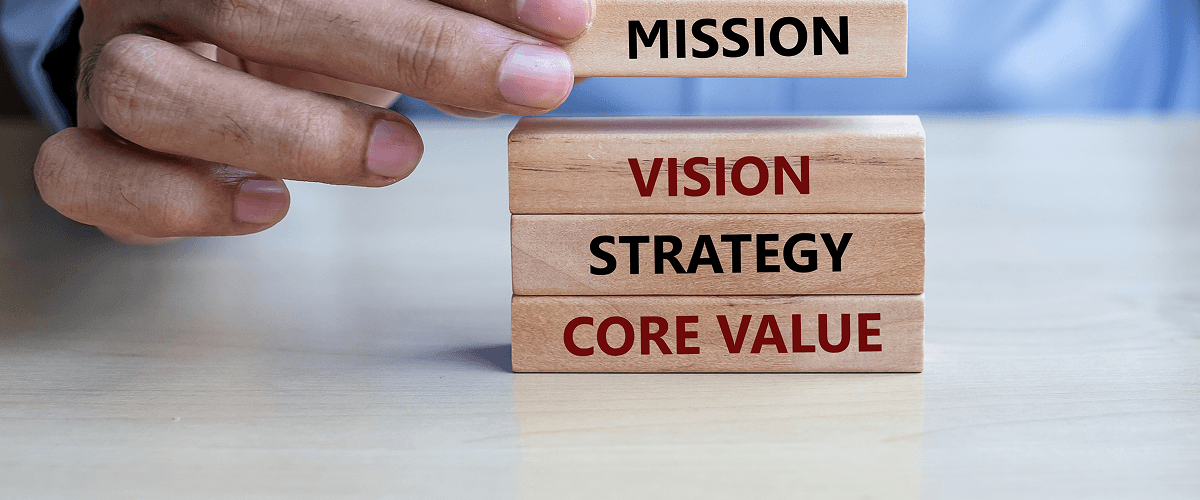 A hand placing a wooden block labeled "MISSION" on a stack of blocks that say "VISION," "STRATEGY," and "CORE VALUE," representing the benefits of communicating a company's values through video.