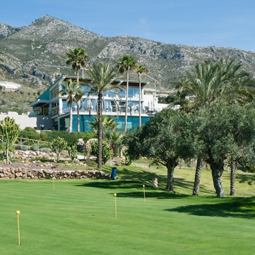 Modern building surrounded by palm trees and greenery, located in a mountainous area with a manicured lawn in the foreground.
