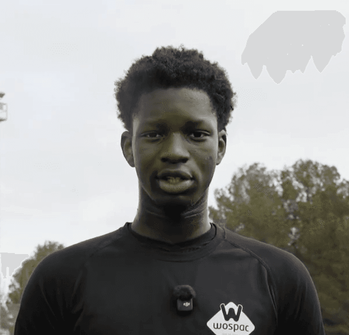 Person in grey sweater and blue shirt facing camera with neutral expression against light grey background