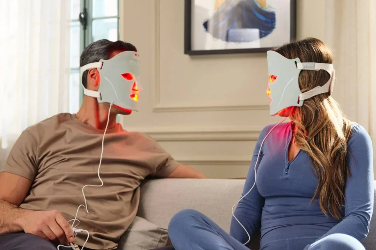 A man and a woman sitting on a couch wearing LED light therapy masks facing each other.