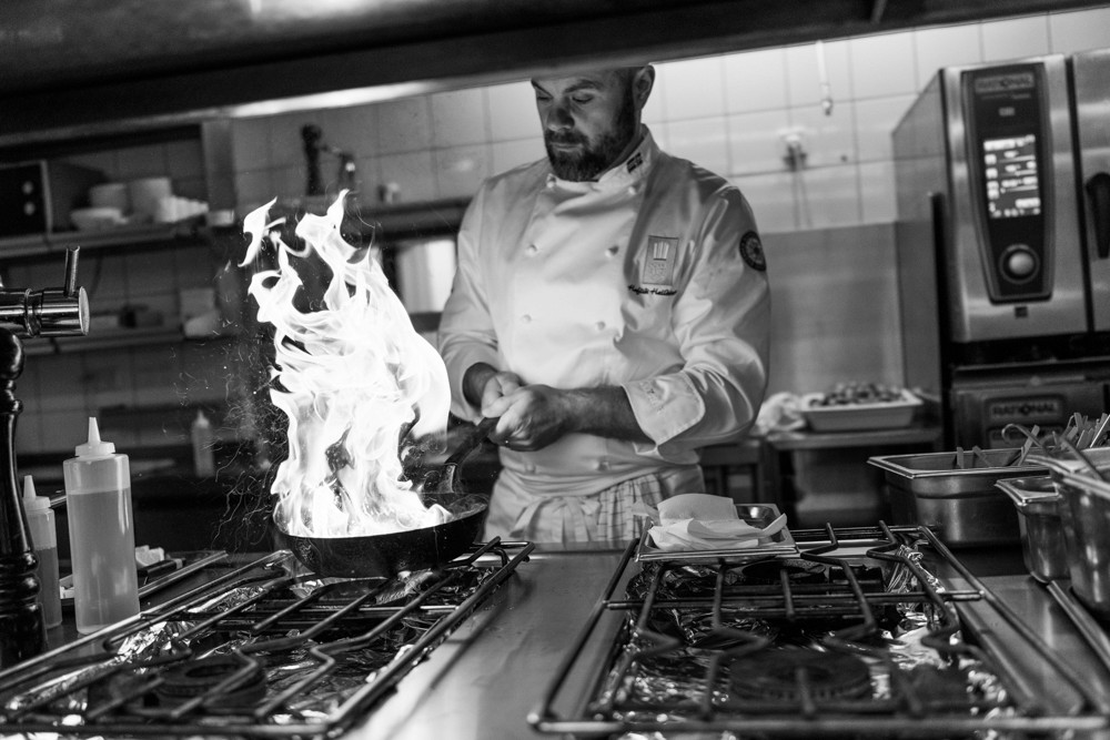  Icelandic chef Ísak in a tall white toque plates food in a professional kitchen under warm hanging lights.