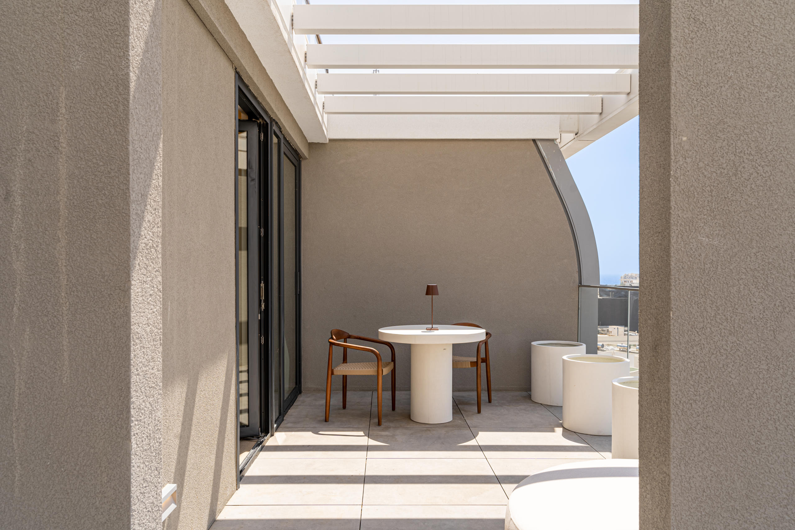 A modern outdoor space featuring a table, two chairs, and sleek architectural elements under a shaded roof.