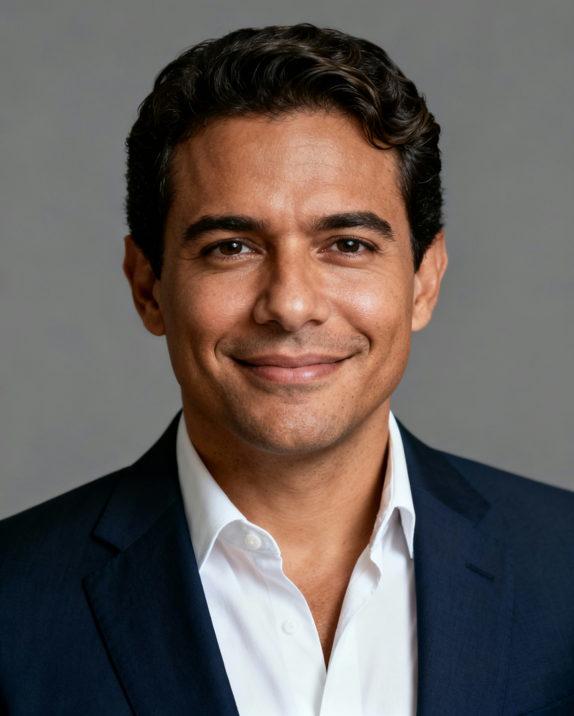 A professional portrait of a man in a navy suit and white shirt, smiling warmly against a neutral gray background.