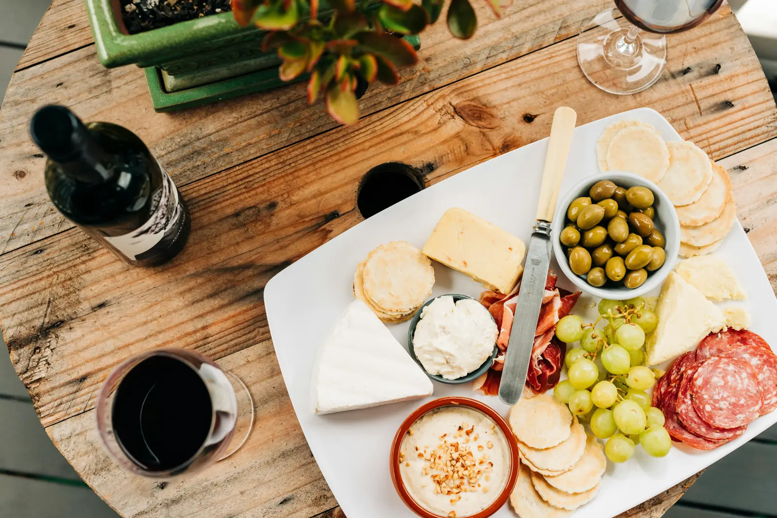 A stylish wooden table features a charcuterie board with assorted cheeses, cured meats, green olives, grapes, and crackers, alongside a bottle of red wine and two glasses, enhanced by a potted succulent centerpiece.