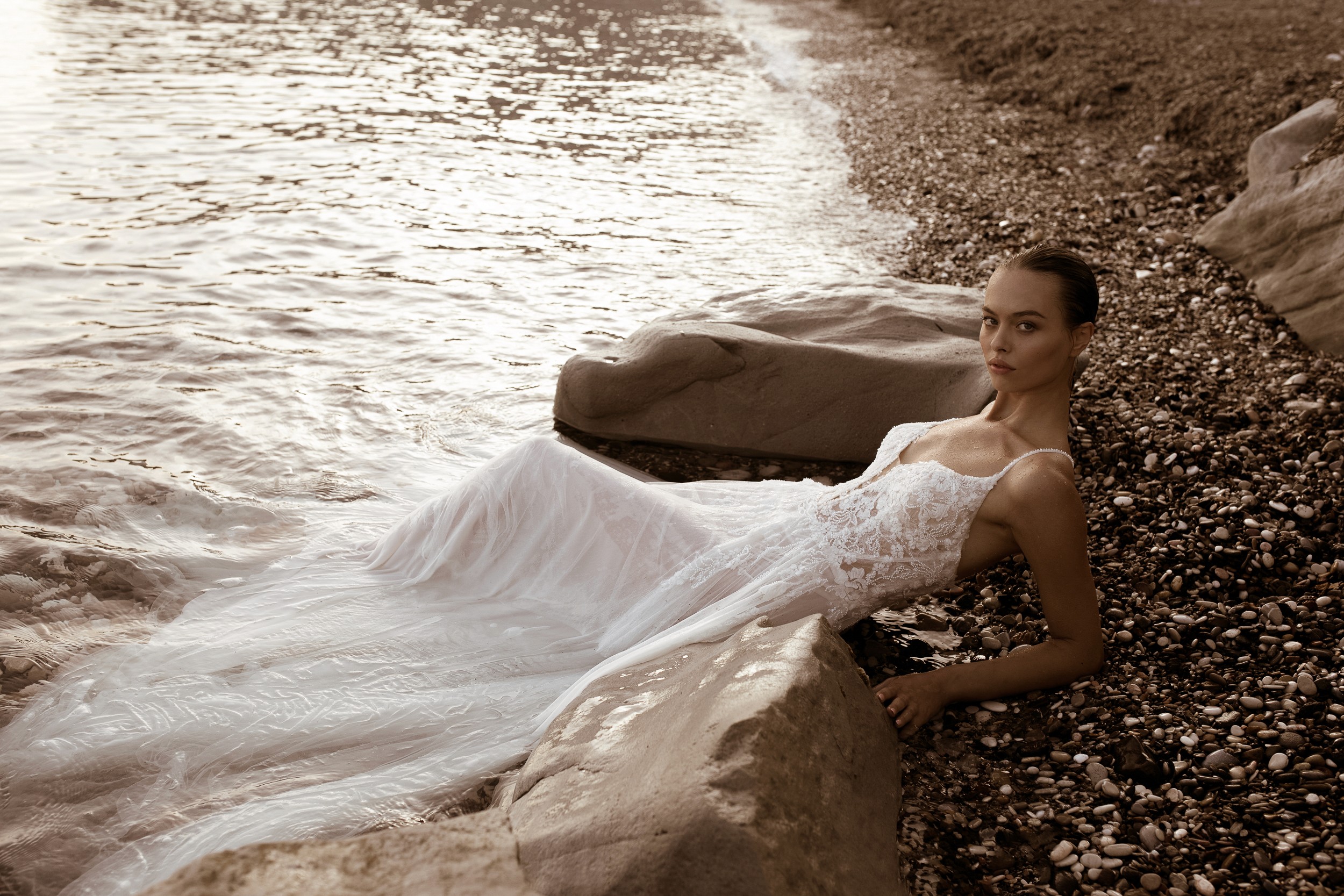 Ein elegantes, bodenlanges Brautkleid aus leichtem, fließendem Stoff steht im goldenen Licht des Sonnenuntergangs am Strand. Das Kleid hat eine romantische, luftige Silhouette, die perfekt zur entspannten Atmosphäre der Hochzeit am Meer passt. Im Hintergrund zeichnet sich der Horizont mit einer malerischen Farbpalette ab, während das Brautpaar den besonderen Moment genießt.