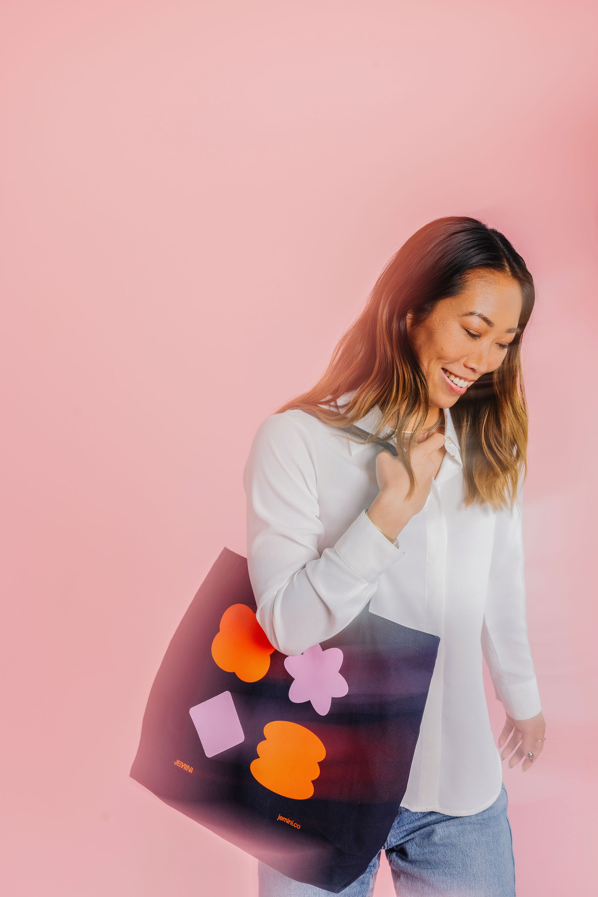 Woman smiling while carrying a tote bag with colorful geometric shapes against a soft pink background.