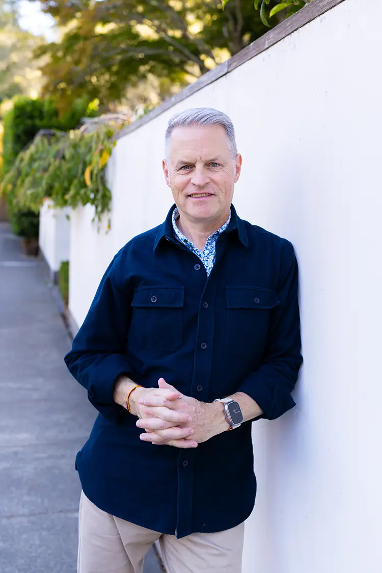 John Coyne, PsyD, a therapist in Marin specializing in chronic illness and insomnia, is leaning against a white wall outdoors