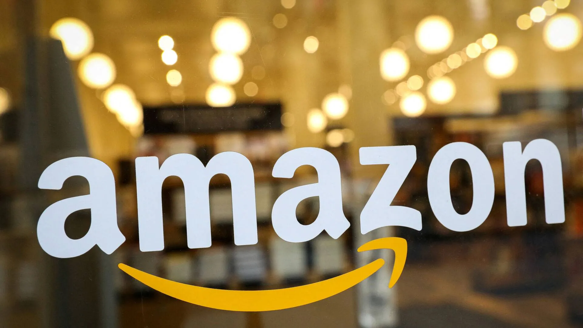 Amazon logo displayed on a glass window, with warm glowing lights blurred in the background, highlighting the company's branding in a retail setting.