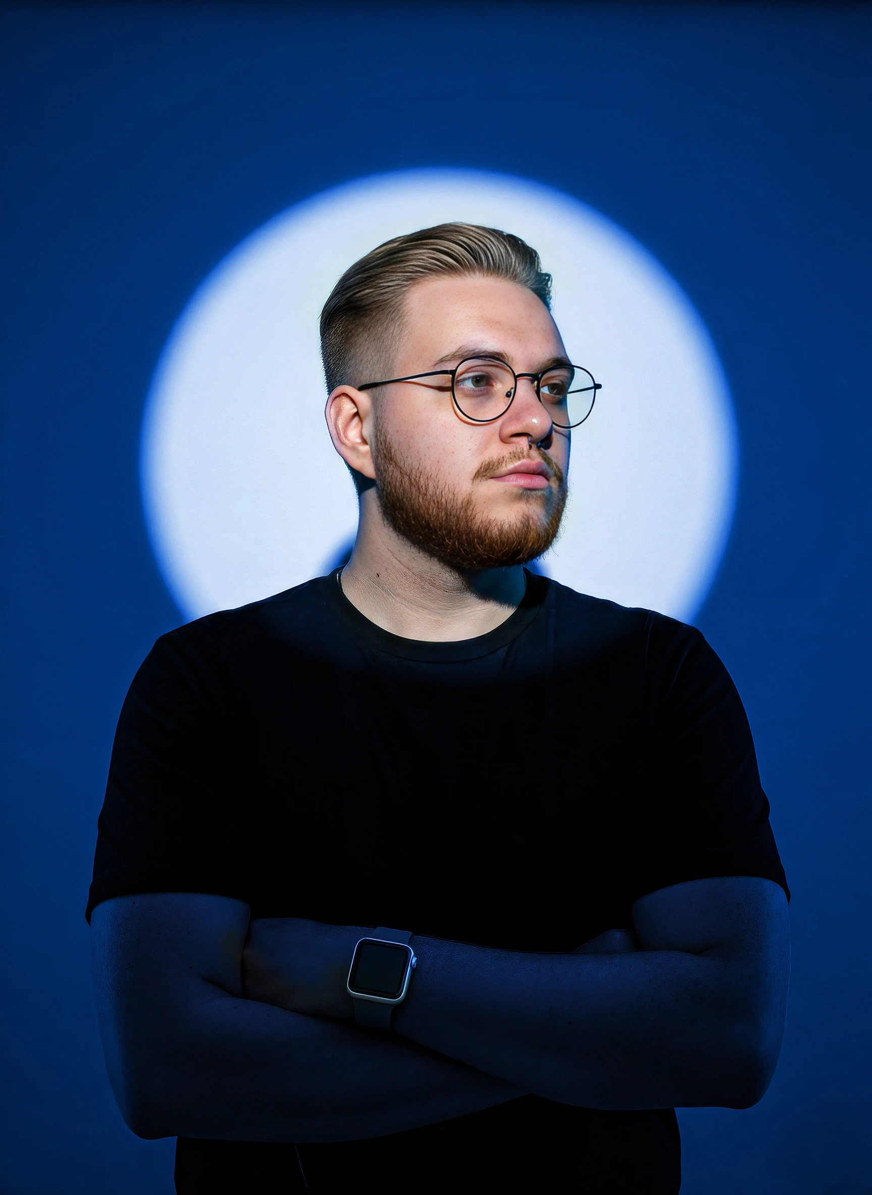 A young man with round glasses, a nose ring, and facial hair looks toward the camera against a dark, softly colored background.