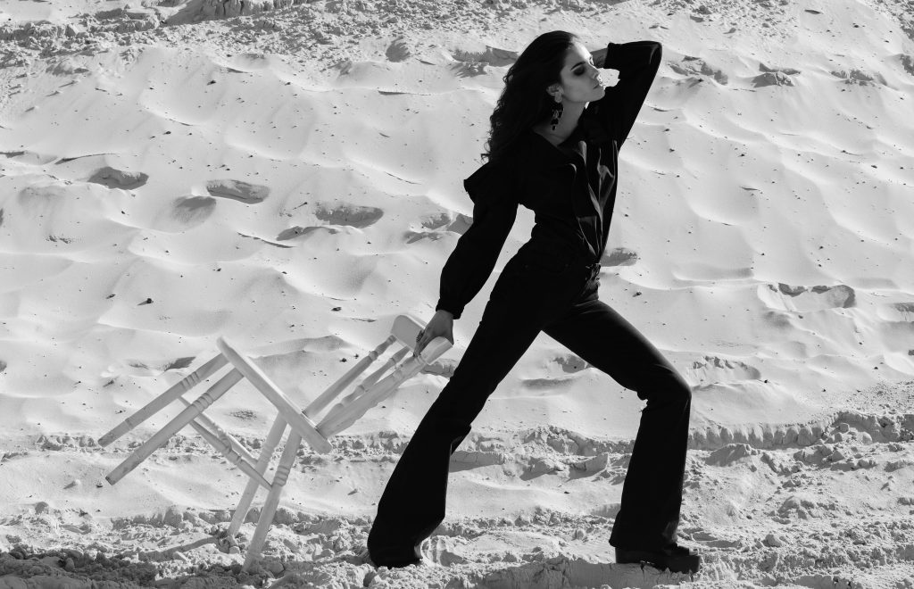 Zwart-witte modefoto van een model in het zwart dat op het zand poseert met een witte stoel in de hand