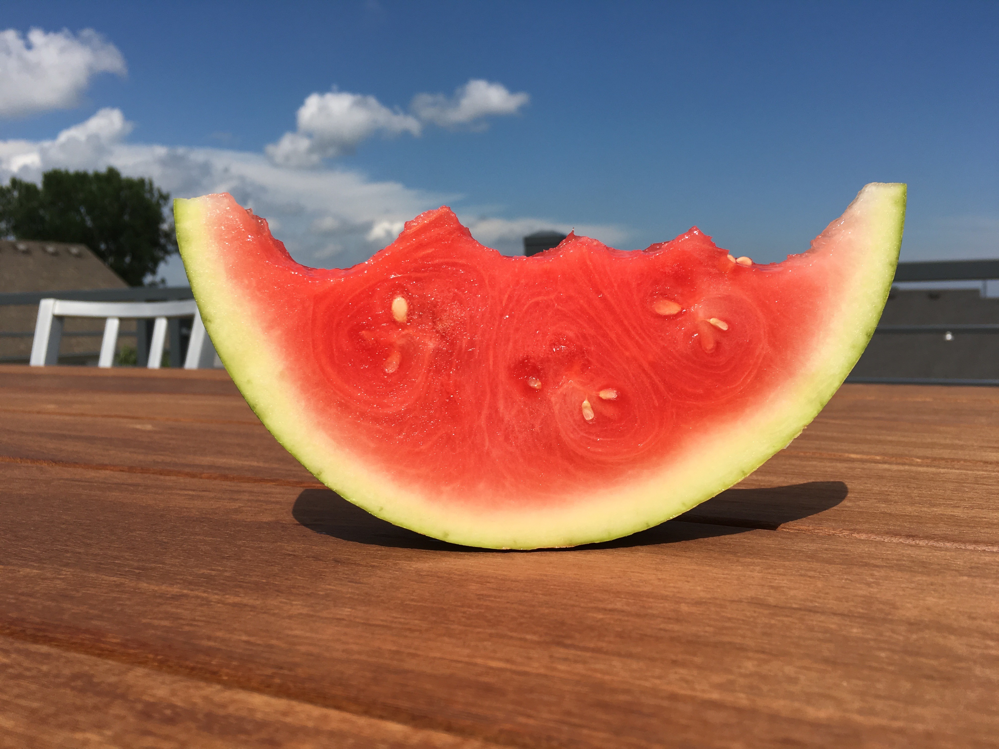 A slice of watermelon rests on a wooden table, with bright red flesh and green rind against a blue sky.