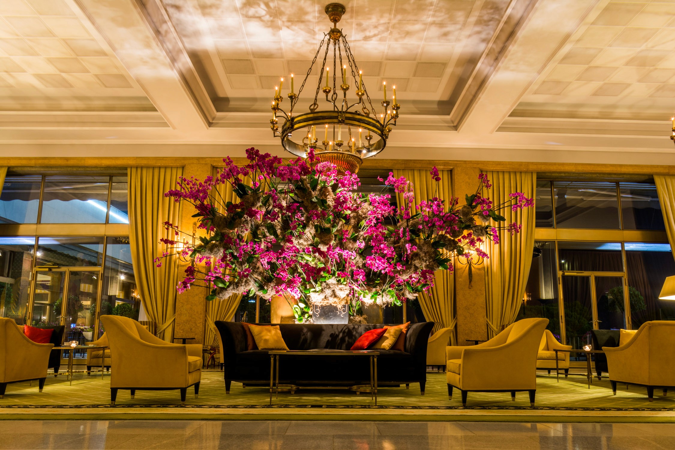 A luxury hotel lobby features a large floral arrangement of pink and purple flowers under a crystal chandelier.