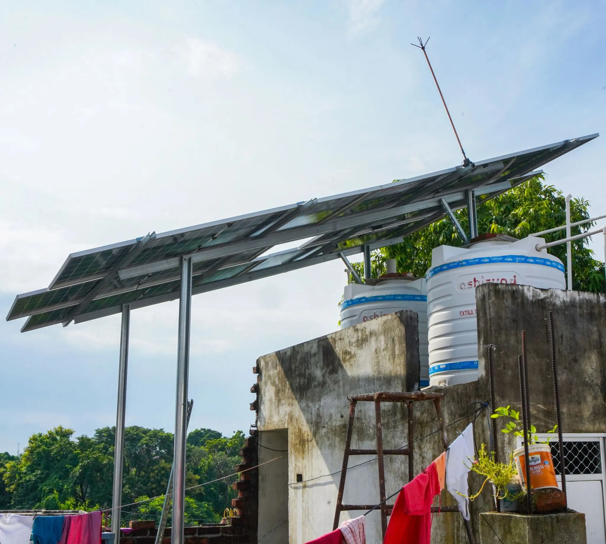 Elevated solar panel structure on a house rooftop next to a water tank and potted plants