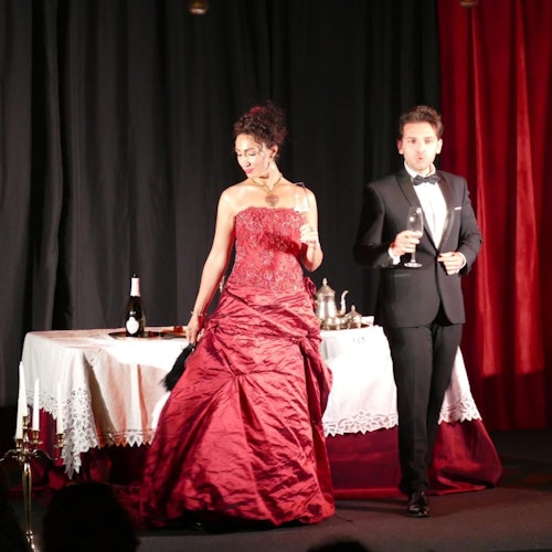 Une femme en robe rouge et un homme en smoking tenant un verre se tiennent près d'une table avec une nappe blanche, une bouteille et un service à thé.