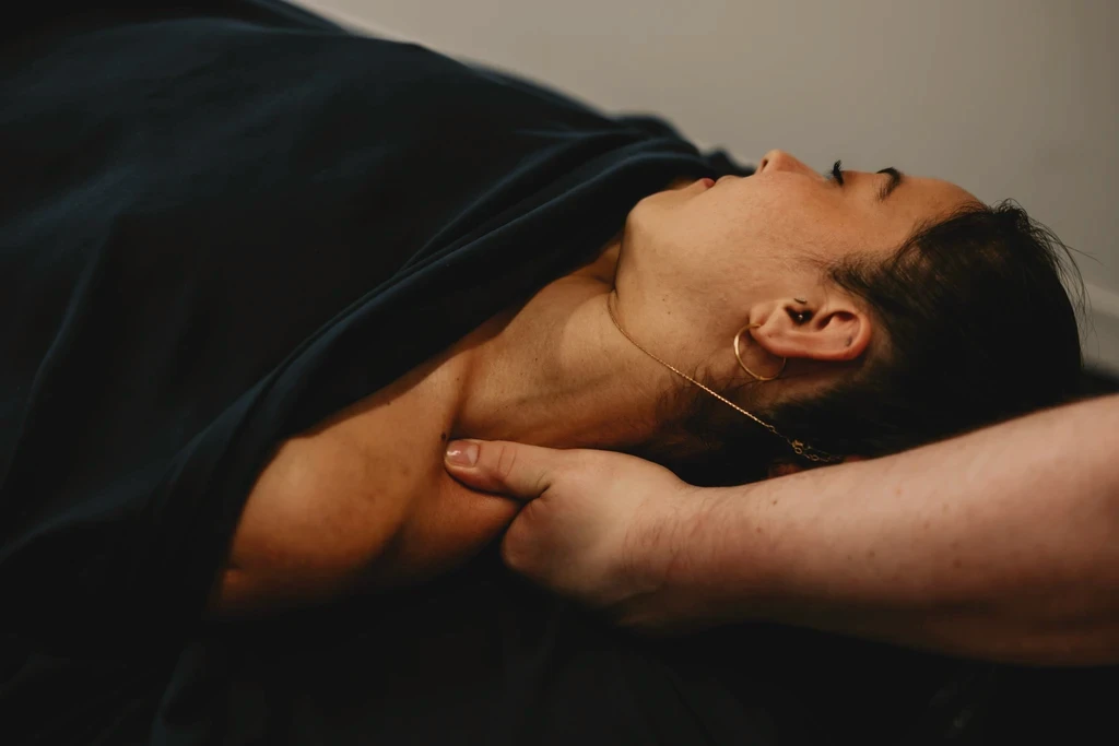 A massage therapist applying deep tissue pressure to a person's neck and shoulder muscles while lying face up on a massage table.