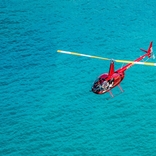 เฮลิคอปเตอร์สีแดงพร้อมใบพัดปลายสีเหลืองบินเหนือผืนน้ำสีฟ้าครามที่สดใส