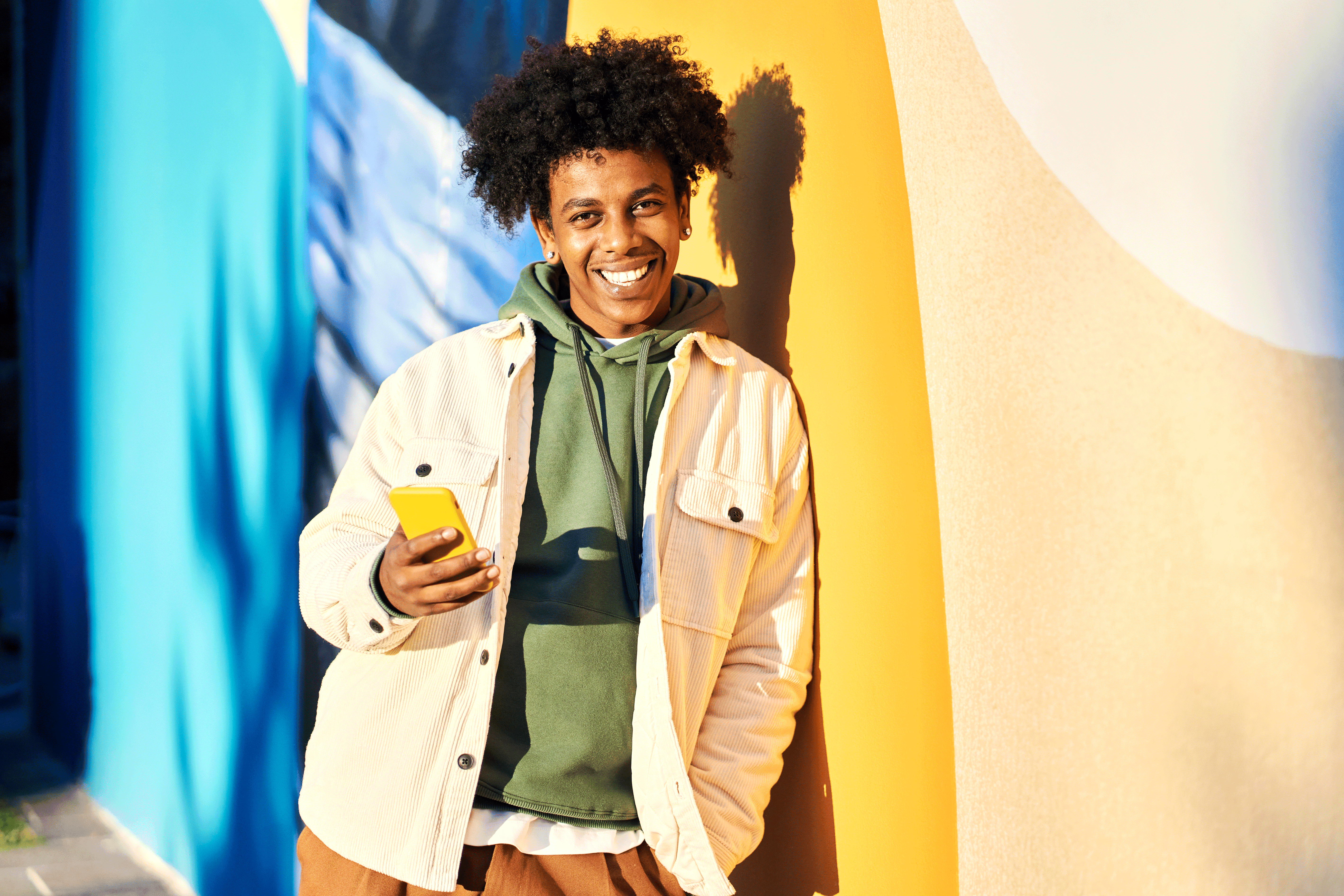 A smiling person with curly hair leans against a vibrant, sunlit mural while holding a yellow smartphone. They are wearing a green hoodie layered under a cream corduroy jacket.