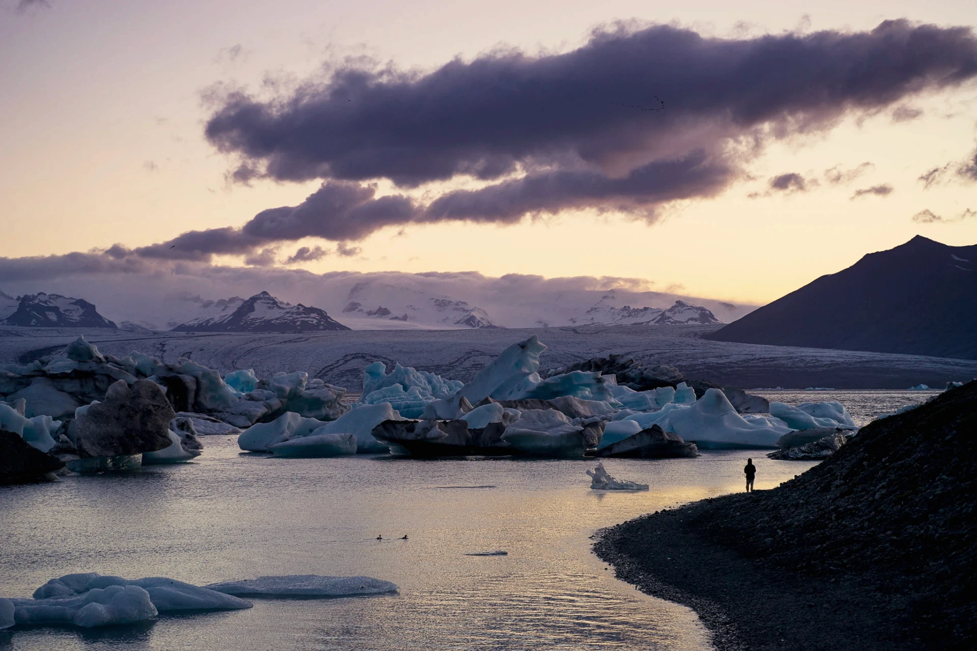 Person standing by a glacial lagoon with icebergs at sunset.