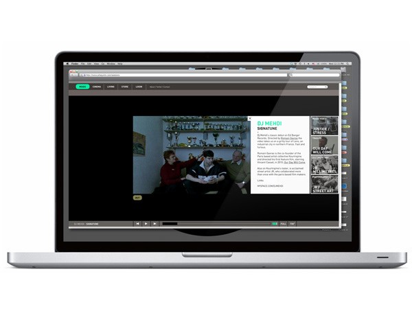 A silver Apple MacBook laptop is open, displaying a webpage with a black background and white text. The webpage features a photo of a man and a woman seated at a table, with text including “DJ MEHOI” and “SIGNATURE”. The browser’s address bar shows “www.djmehoi.com”. The laptop’s screen is angled slightly to the right.