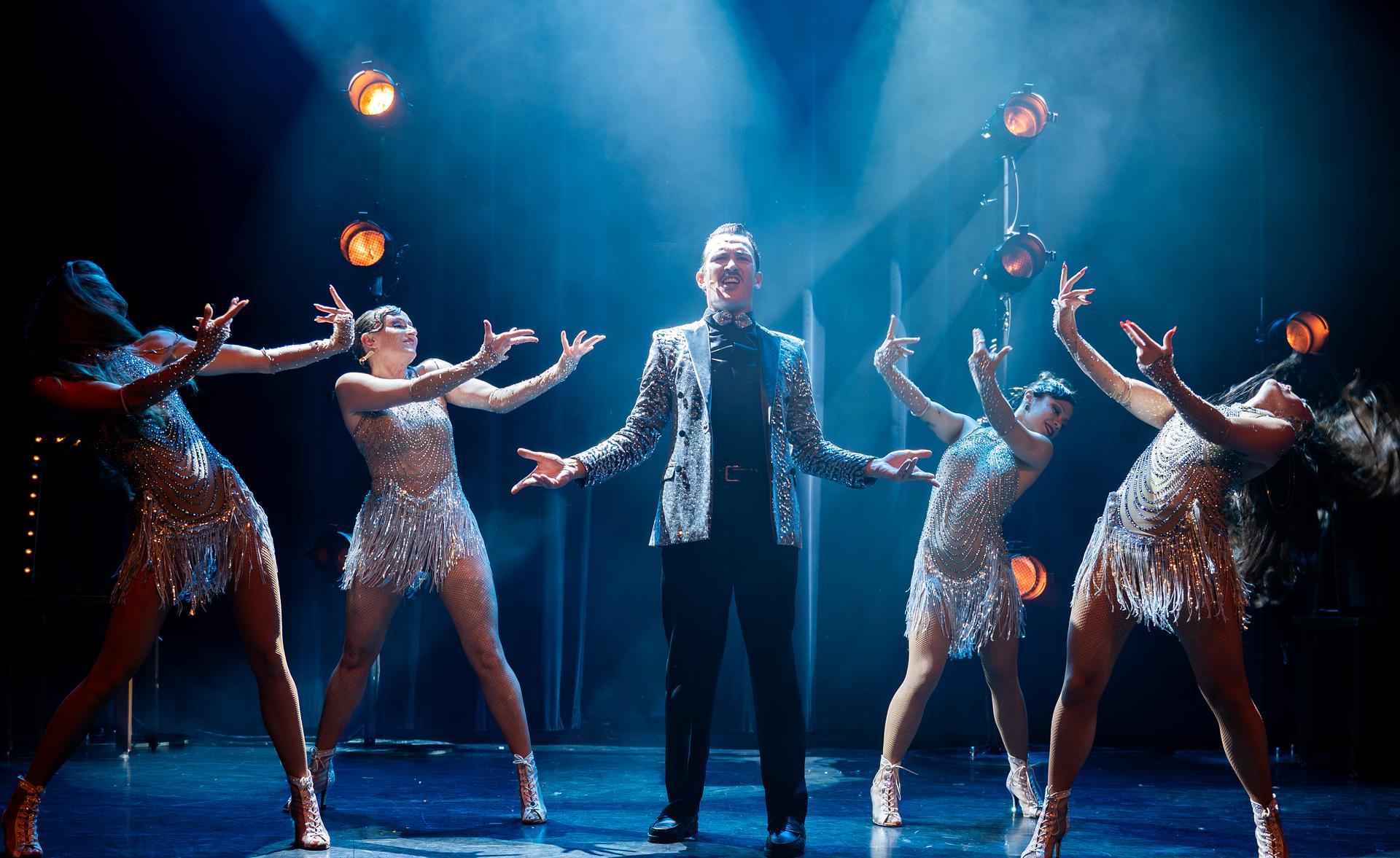 Sur scène, James Warner en veste à sequins argentés, les bras ouverts, entouré de ses quatre muses en robes à franges, sous les faisceaux bleus des projecteurs