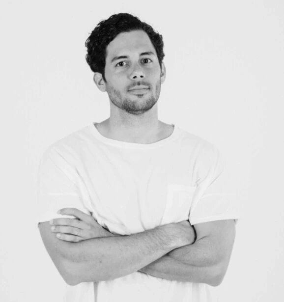 Portrait of a man with curly hair, wearing a white t-shirt and standing with his arms crossed against a plain background