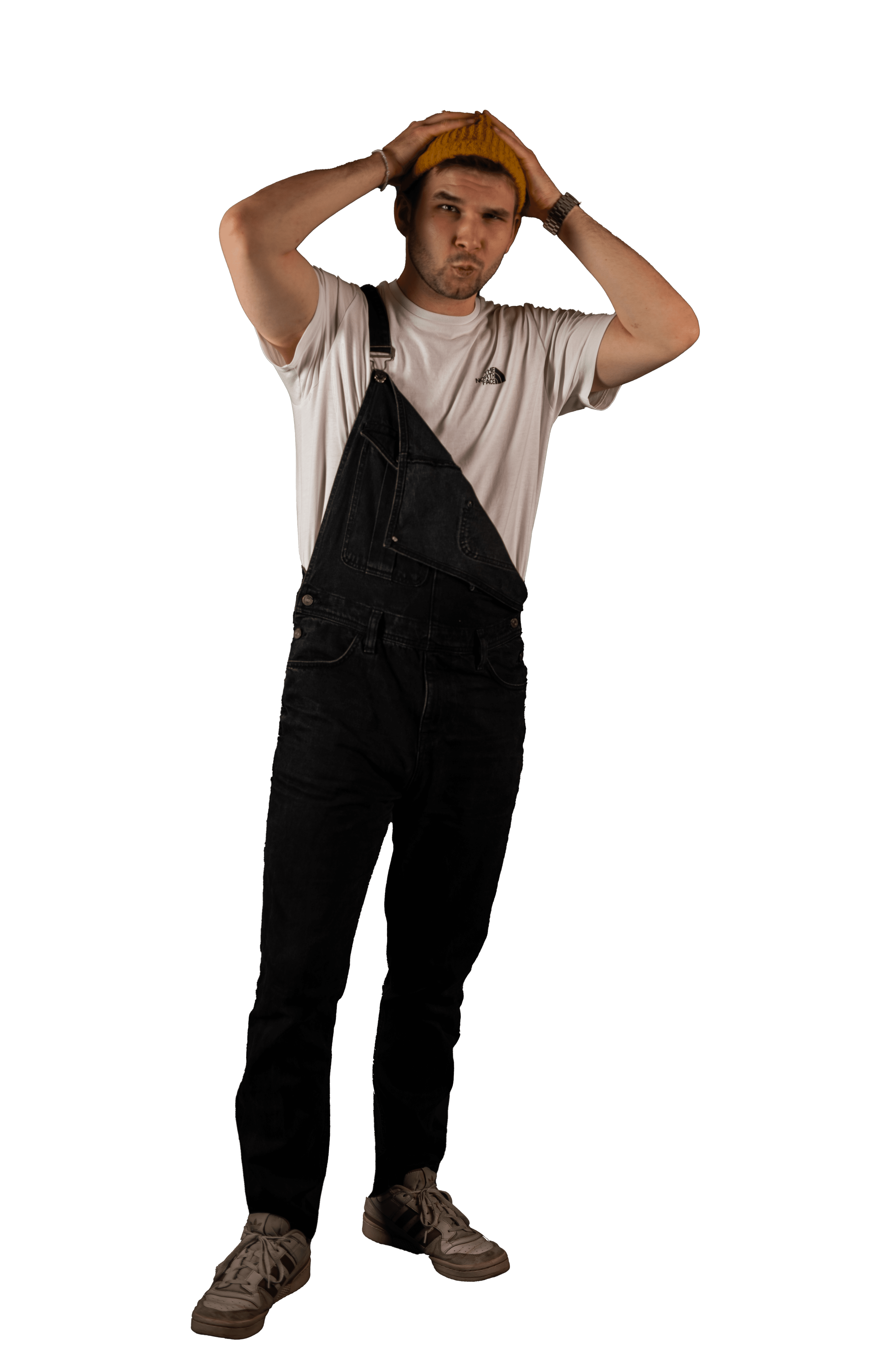 Ganzkörperaufnahme des Musikers Lasse PTS. Er trägt eine dunkle Latzhose über einem weißen T-Shirt und eine gelbe Mütze, die er mit beiden Händen anpasst, während er direkt in die Kamera schaut.