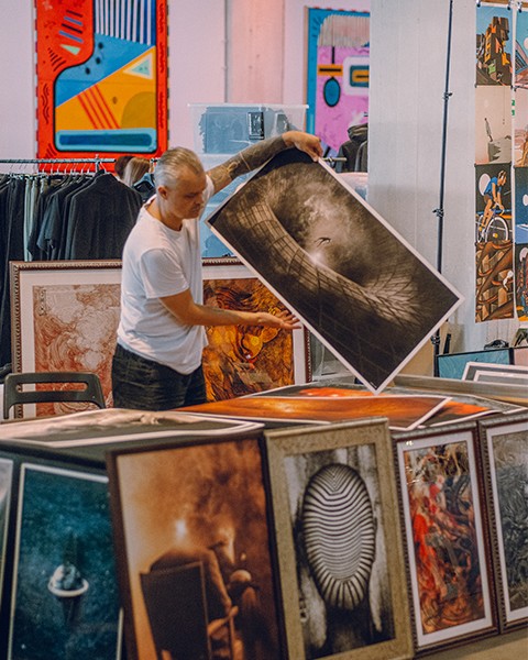 Zdjęcie przedstawiające artystę, który porządkuje swoje plakaty i obrazy na stanowisku targowym.