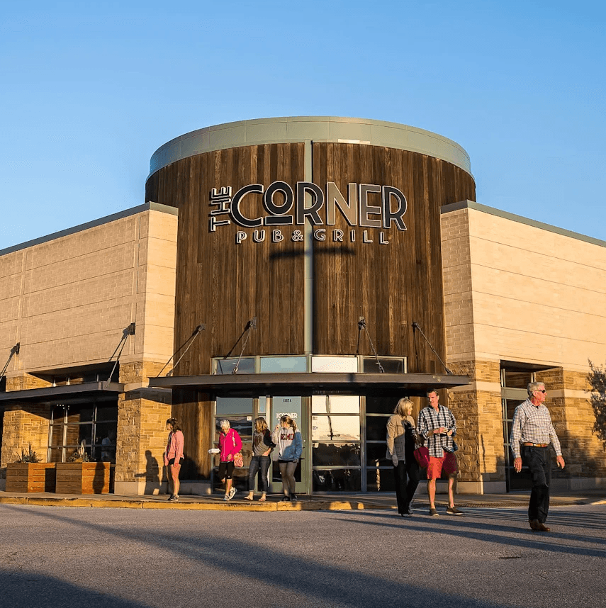 Exterior view of a shopping mall entrance with people walking in front, under a clear blue sky.