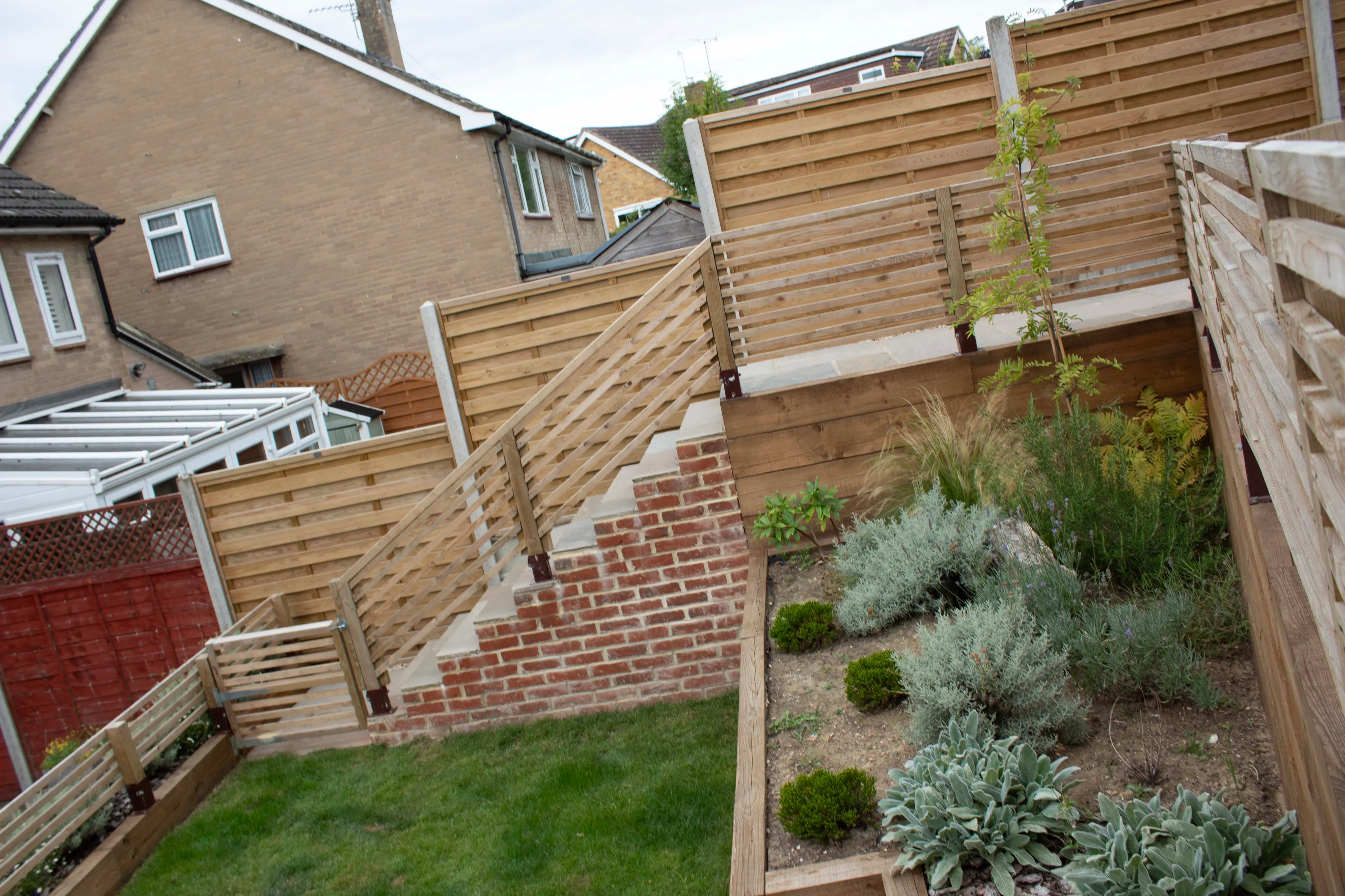 A garden featuring wooden fencing, steps leading up a slope, and green shrubs in landscaped beds.