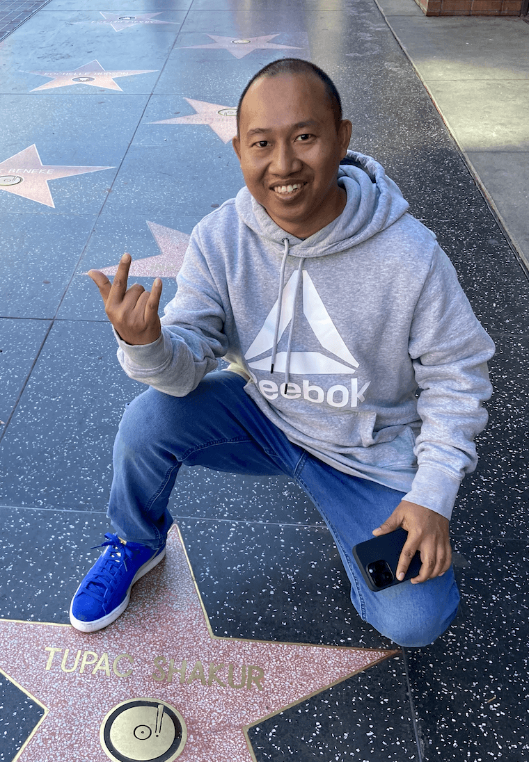 A person stands outdoors in a natural setting with lush green trees under a clear sky, wearing a dark sweatshirt, and looking into the distance.