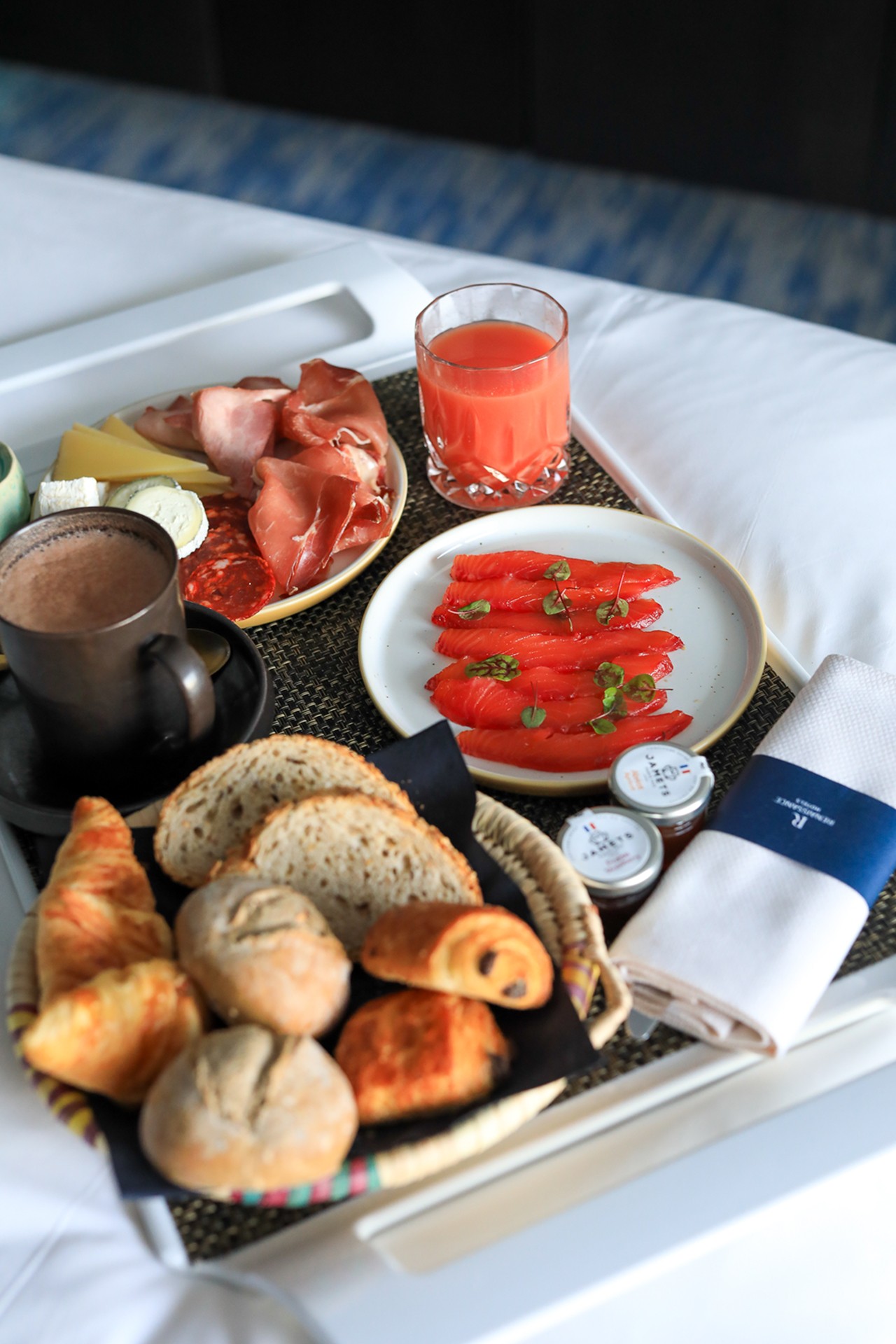 Un plateau de petits-déjeuners présente des tranches de saumon fumé garnies de câpres, accompagnées de charcuterie, de pains assortis, de viennoiseries et d’un verre de jus d’orange, mettant en avant un cadre de service en chambre de luxe dans un hôtel.