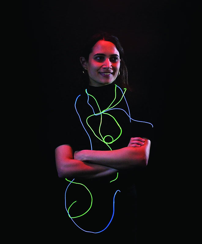 A person with dark hair stands with their arms crossed against a black background. Behind them, a neon green and blue neon line design forms the shape of a person’s torso and arms. The person is smiling and looking directly at the camera.