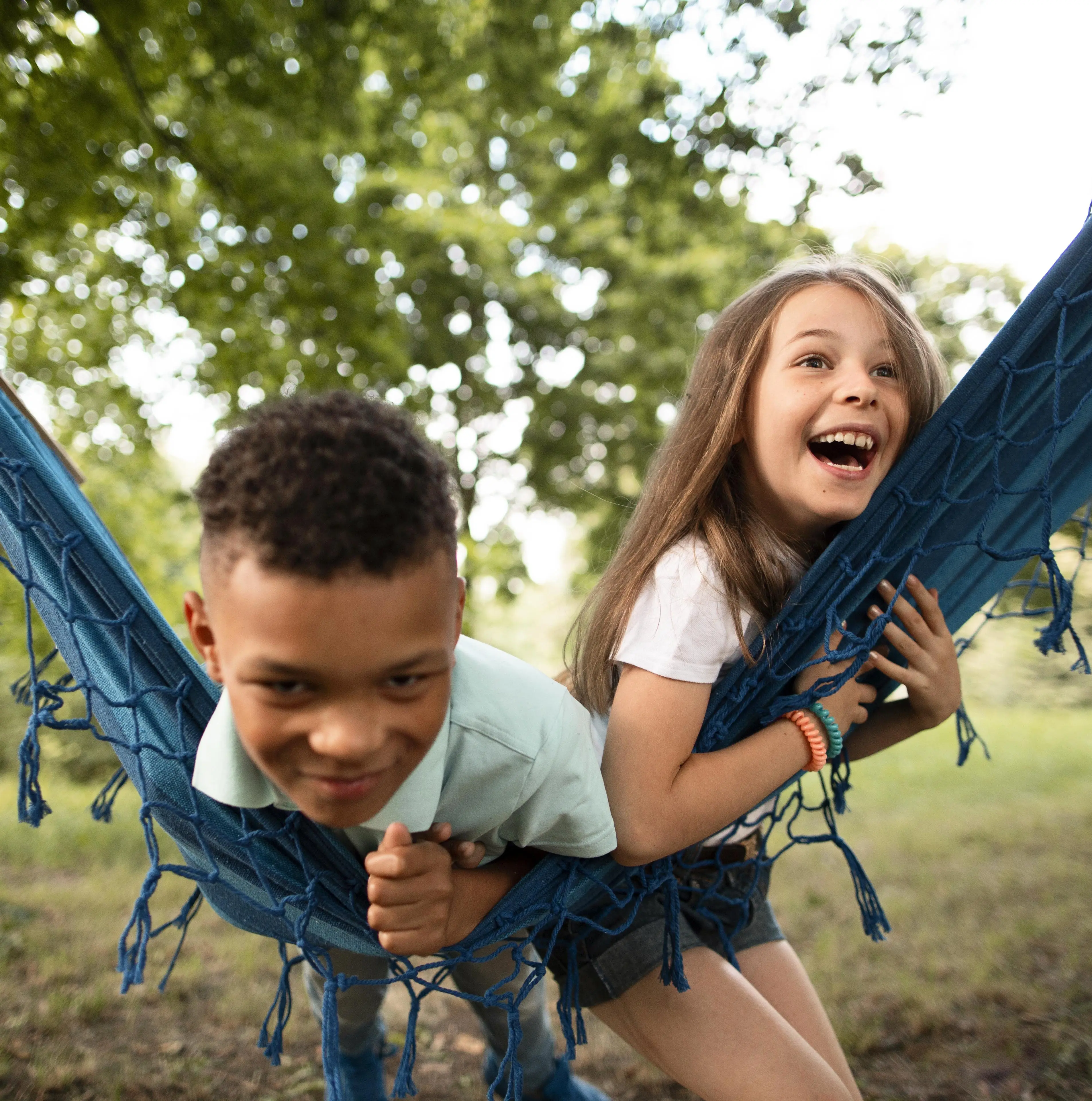 Foto de duas crianças, um menino e uma menina, deitadas em uma rede azul ao ar livre em um parque ou jardim, ambas sorrindo e olhando para cima. A menina tem cabelo castanho comprido e o menino tem cabelo escuro e cacheado.