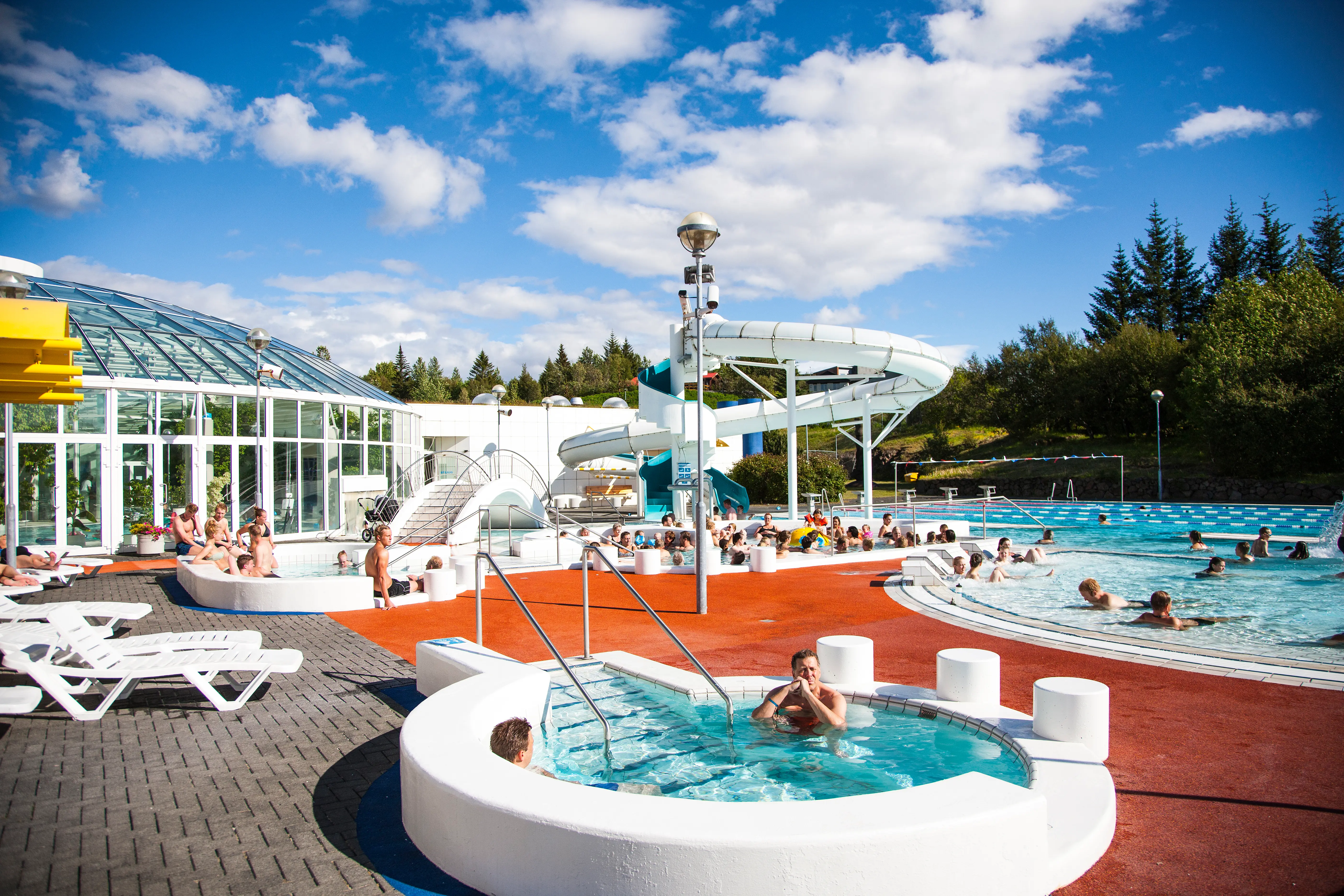 Families and swimmers enjoying the warm outdoor pools at Árbæjarlaug in Reykjavík on a sunny day, with kids’ play areas, hot tubs, and a large waterslide visible, reflecting Iceland’s year-round geothermal pool culture.