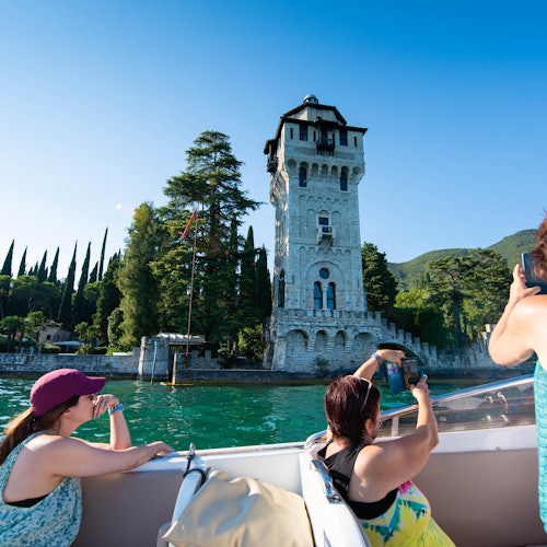 Three people on a boat taking photos of a stone tower by a lake, surrounded by trees, with a clear blue sky in the background.