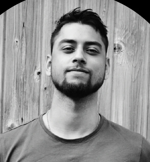 A man with a short beard and dark hair stands against a wooden background, looking confidently at the camera.