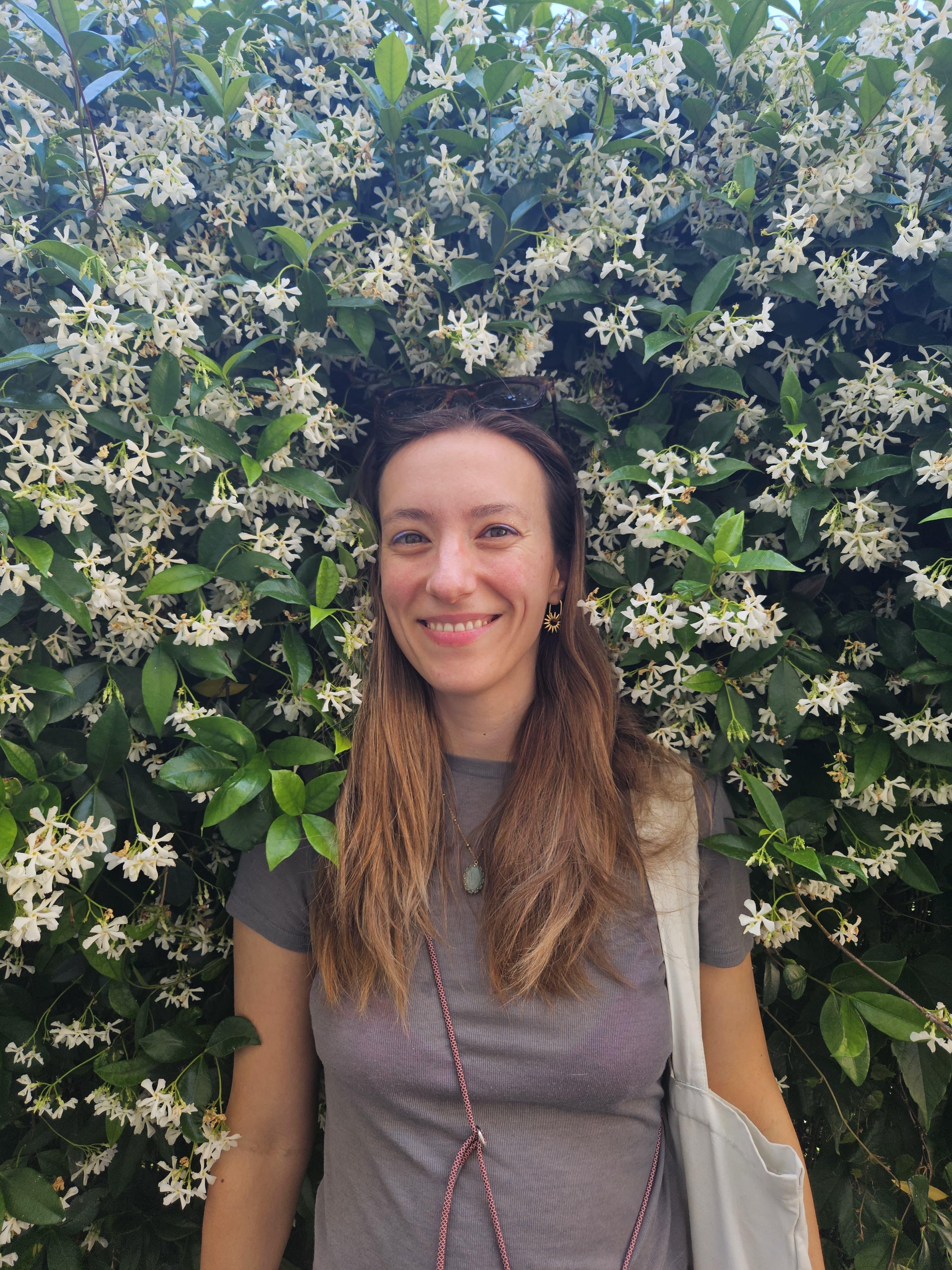 A woman with long brown hair stands smiling in front of a lush hedge covered in blooming white star-shaped flowers, wearing a gray top and carrying a light-colored bag, surrounded by vibrant green leaves.