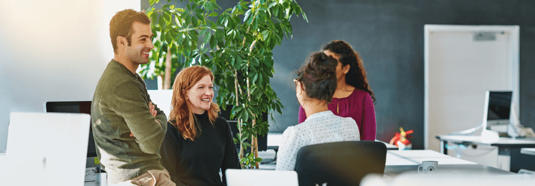A group of people engage in discussion in a modern office setting, surrounded by plants and a sleek design.