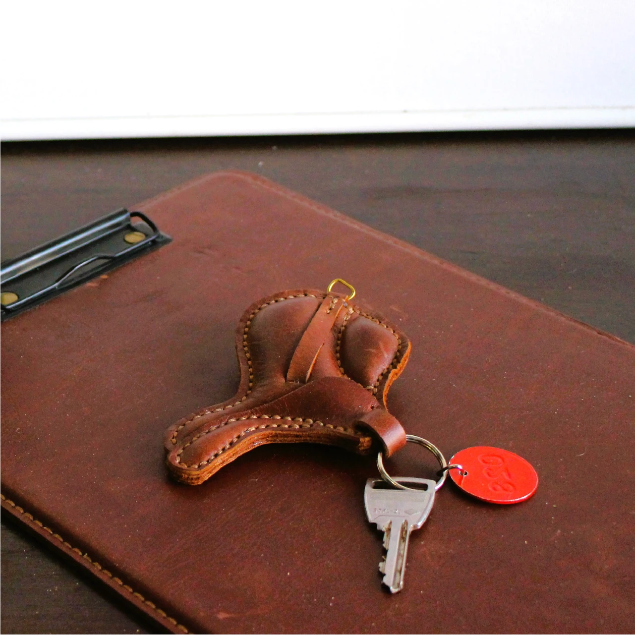 Brown leather keychain on a leather folder, with two keys and a red tag. The scene conveys a rustic, minimalist feel.