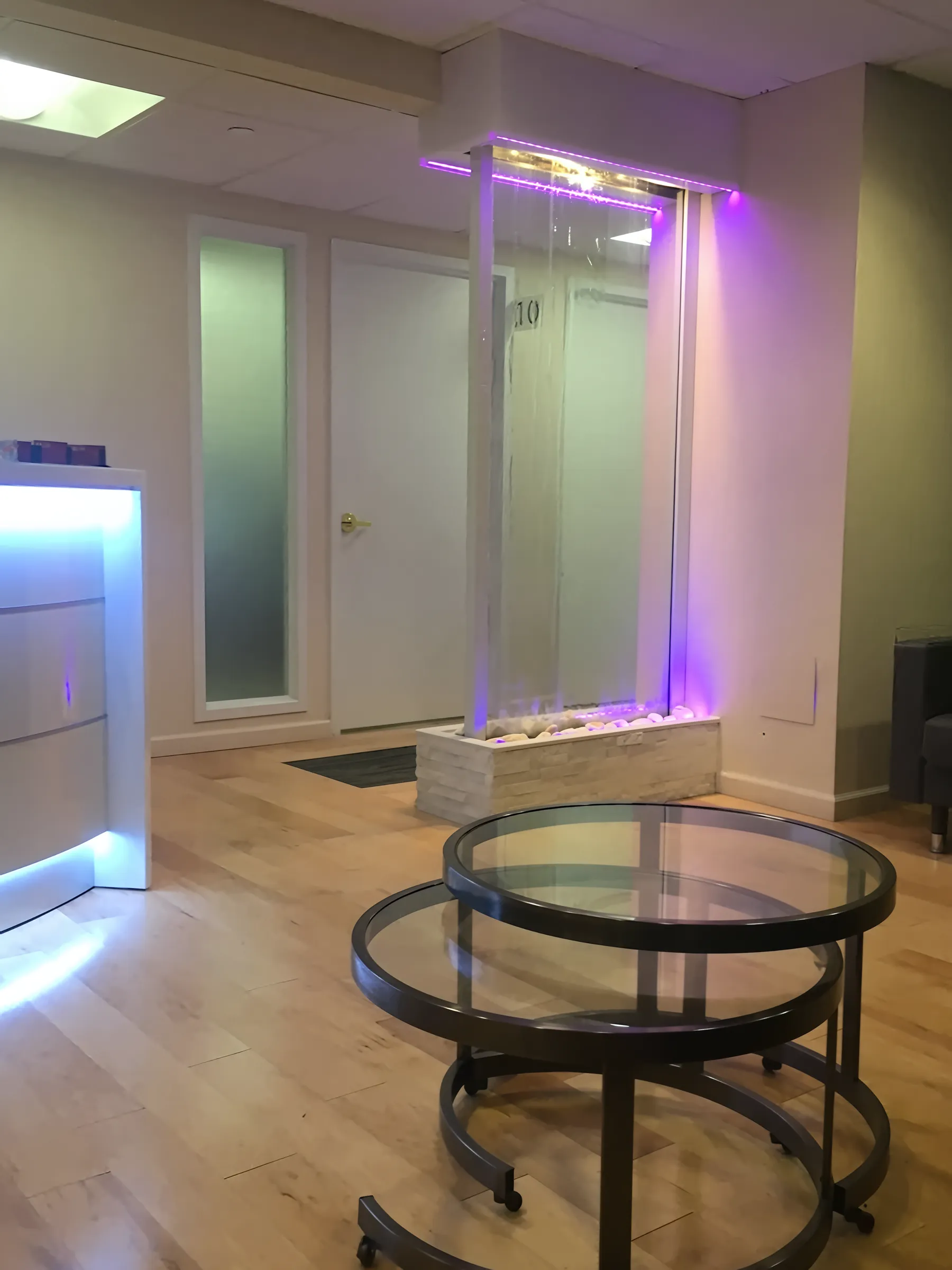 Hardwood flooring in a Stoll Medical Group interior hallway with a decorative LED-lit water feature, frosted glass doors, and glass table.